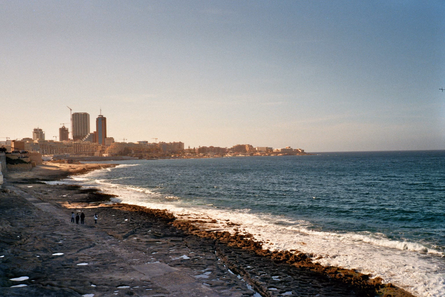 Felsige Küstenlinie auf Malta mit Wellengang und einer modernen Stadtsilhouette im Hintergrund.