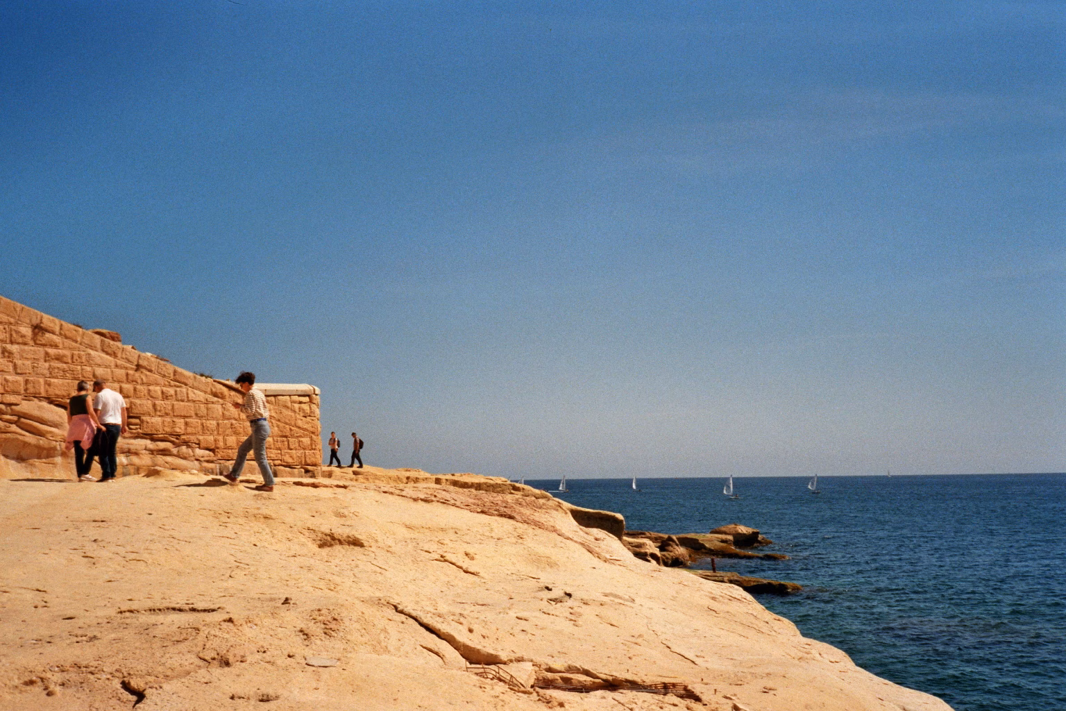 Spaziergänger auf den hellen Kalksteinklippen Maltas mit Blick auf das blaue Meer und Segelboote.